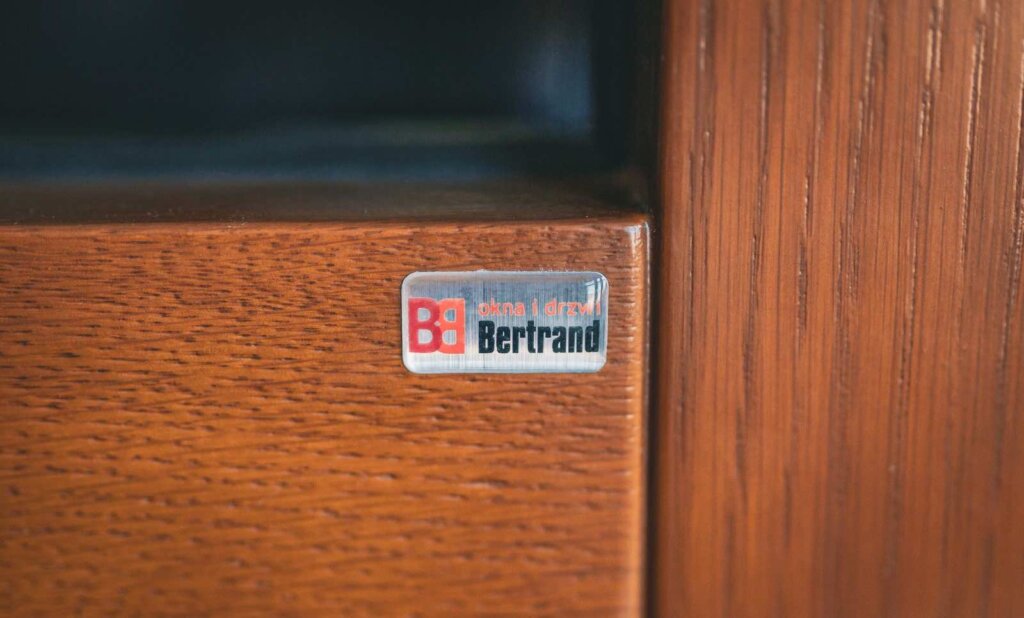 srebrna naklejka 3d z napisem Bertrand na brązowej ramie okiennej